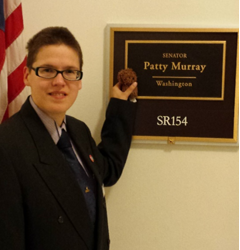 picture is of person(me) wearing black double buttoned suit standing in front of Patty Murray Office in Washington DC. I am holding a small brown stuffed Chewbacca. I call him chewy. He my comfort buddy. I am looking at camera. I have short dark brown hair. 